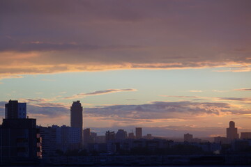 A beautiful cityscape with silhouettes of tall buildings stretching into the distance against the background of a blue sky and gray-yellow clouds from the setting sun