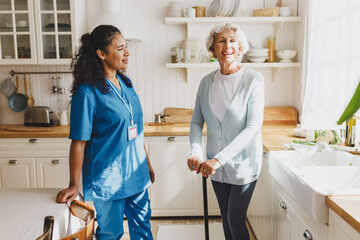 Happy smiling elderly woman using walking stick after listening instructions of its usage from pretty black female medical nurse standing next to her in kitchen. Medical assistance at home - obrazy, fototapety, plakaty
