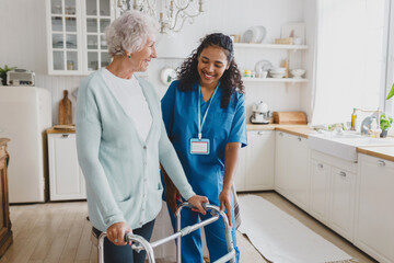 African american medical nurse volunteer helping senior female at home, teaching her to walk with walker, smiling and giving instructions how to use device, standing together in cozy kitchen - 673354857