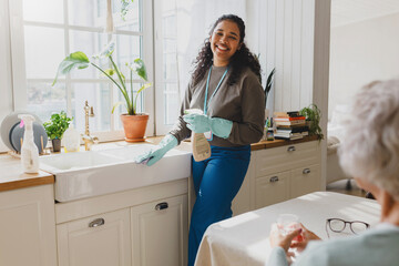 Fototapeta Selective focus on happy african american volunteer helping senior lady to clean house, standing in kitchen in raisin gloves washing sink with detergent, talking to elderly female sitting at table obraz