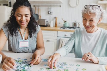 Portrait of two people at kitchen table, african american female volunteer spending leisure time with senior lady, playing together puzzle game, chatting and having fun. Social support - 673355055