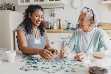 Cheerful senior lady having fun plying puzzle game at kitchen table with black female volunteer, taking care of her as social support of retired persons, spending leisure time, talking - 673355070