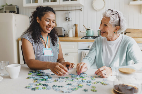 Cheerful senior lady having fun plying puzzle game at kitchen table with black female volunteer, taking care of her as social support of retired persons, spending leisure time, talking