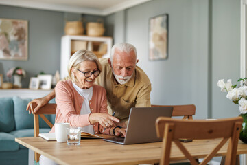 Senior couple sit at a table in the living room with laptop,working from home office - 673363670