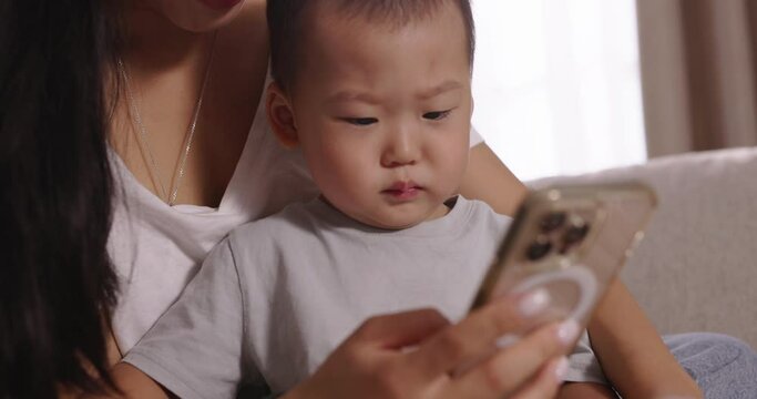 Mom and child on sofa looking to smartphone. Cinematic Chinese, Japanese, Korean, Asian. Close up of pretty young mother spending time with little child and browsing on cell phone, using mobile phone