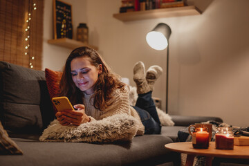 A young woman spends a cozy winter evening at home lying on the sofa looking at her smartphone. Winter holidays, Christmas and online surfing concept - 673372031