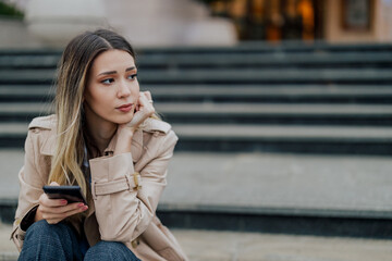 Worried young stylish female sitting on a stairs outside and using a phone