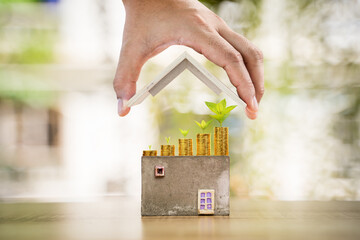 Woman hand hold to open a roof and a coin stack substantial savings money inside the house and plant growing on the top in the public park, Business investment or loan for real estate concept.