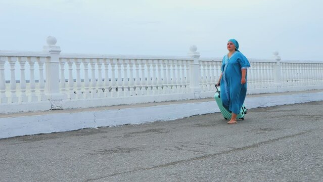 A woman in a turquoise dress and headscarf walks early in the morning with a suitcase along the embankment against the backdrop of the sea in the wind. Overall plan