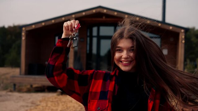 A girl holds a bunch of keys to her new home against the backdrop of wooden cottage in the woods in the countryside
