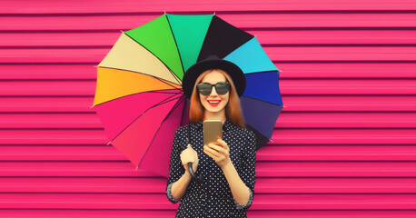 Fototapeta premium Portrait of happy smiling young woman looking at mobile phone with colorful umbrella on pink background