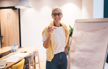 Cheerful senior female entrepreneur standing near flip chart with graph