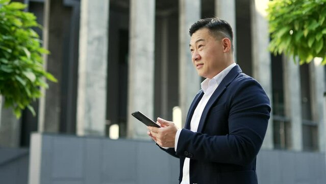 Side view of asian businessman in formal suit looking at mobile phone walking in street to office beside green pine trees. Entrepreneur browsing scrolling smartphone, chatting online or checking email