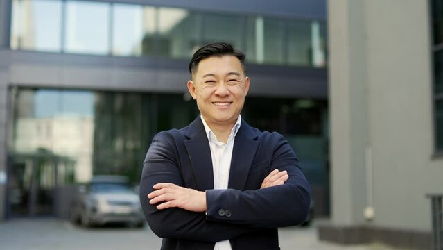 Portrait of a happy asian businessman in formal suit standing on the street near office building. Smiling friendly male entrepreneur posing with crossed arms looking at camera. Head shot of the owner
