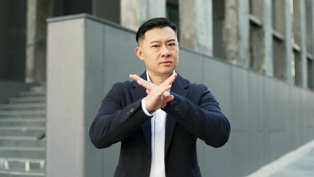 Asian businessman in formal suit showing crossed arms, X sign, shaking head no while standing on street near office building. A serious man demonstrates a gesture of prohibition, denial, disagreement