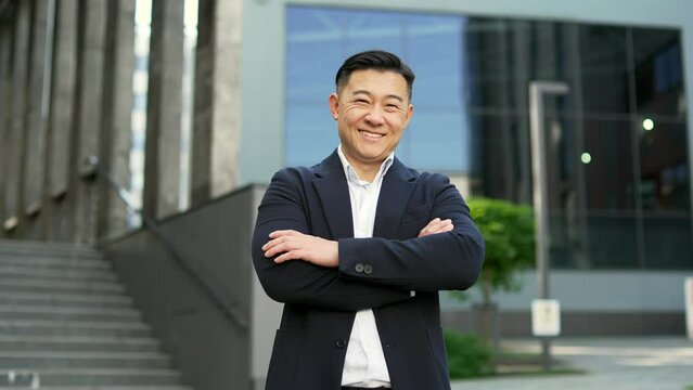 Portrait of a happy asian businessman in formal suit standing on the street near office building. Smiling friendly male entrepreneur posing with crossed arms looking at camera. Head shot of the owner