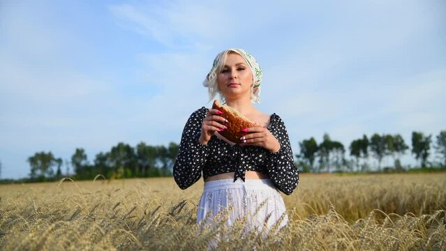 Portrait of sexy blond haired plus size caucasian woman in black blouse with cleavage eating loaf of bread on agricultural wheat field in a sunny summer day. Real time video. Food industry theme.