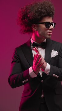 confident fashion best man with sunglasses looking to side, rubbing palms and arranging black tuxedo on colorful background