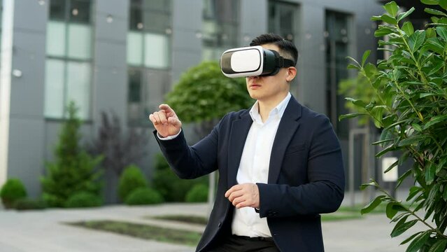 Asian businessman works using VR glasses in virtual reality simulator sitting on a bench on street near office building. Man uses gestures to control computer programs and flips through virtual pages