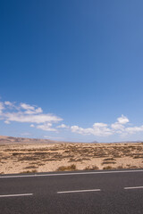 Desert landscape of white sand and desert shrubs. Asphalt road, mountains in the background. Sky with big white clouds. Lanzarote, Canary Islands, Spain.