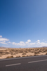 Desert landscape of white sand and desert shrubs. Asphalt road, mountains in the background. Sky with big white clouds. Lanzarote, Canary Islands, Spain.