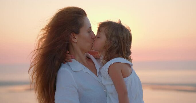 Young mom and little daughter enjoy relaxing at sunset on the ocean. A beautiful mother hugs and kiss child girl tightly. Maternal care and love for the child