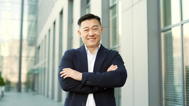 Portrait of a happy asian businessman in formal suit standing on the street near office building. Smiling friendly male entrepreneur posing with crossed arms looking at camera. Head shot of manager