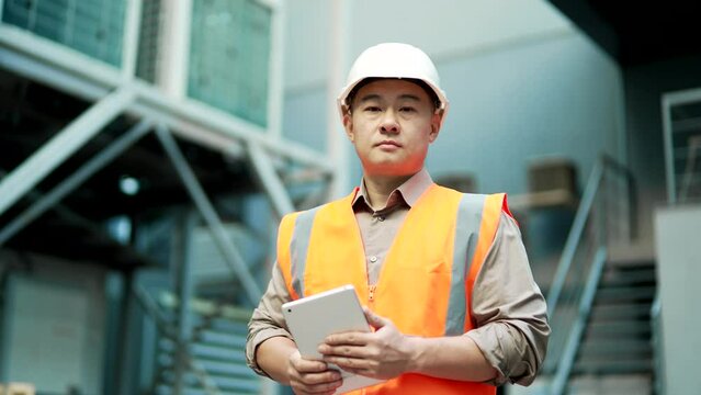 Portrait of confident professional engineer wearing safety helmet and vest standing at factory with digital tablet in hands. Headshot of serious industry manager inspector in uniform looking at camera