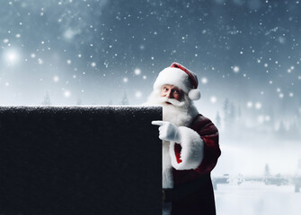 Père Noël souriant en habit traditionnel rouge et blanc,tenant une grande pancarte noire, type tableau noir, et pointant de l'index la zone de texte, sur fond de forêt et village enneigés