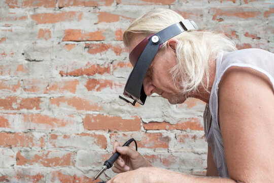 A man with a head loupe and a soldering iron bowed his head over his work, against the backdrop of a brick wall. A device for working with small parts and soldering