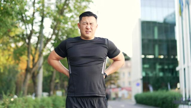 Adult asian runner suffering from back pain, sports injury, muscle spasm in city street. Man in sportswear having back flank ache and problem after exercise jogging outside outdoors in urban city park