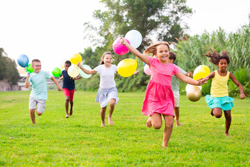 Portrait of cheerful preteen boys and girls with colorful toy balloons in hands running on green lawn in city park on sunny summer day