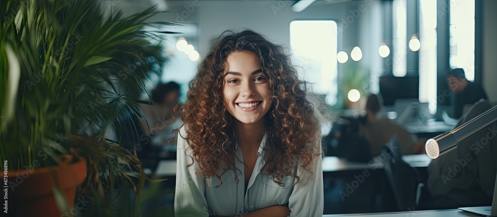 Poster A young female designer situated at her creative workspace in an office is seen smiling towards the camera through a glass