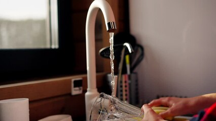 A girl in a checked shirt thoroughly washes dishes with foam in the kitchen of wooden house