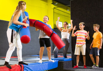 Emotional active preteen girl having funny wrestling by pugil sticks on battle beam with other girl in indoor trampoline arena