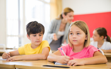 Portrait of focused cute small schoolgirl writing exercises in workbook during lesson in classroom ..