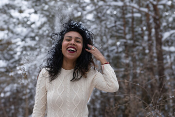 Portrait of Young fashionable stylish brunette pretty latin hispanic woman standing in winter forest or park, falling snow. Smiling winter female portrait outdoors on a winter nature - obrazy, fototapety, plakaty