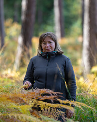 A happy middle-aged woman looks out from behind thick grass and ferns in the autumn forest. Active and joyful lifestyle, life after 50 years