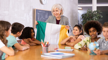 Fototapeta premium Qualified female teacher tells pupils the history of Ireland in lesson and holds the national flag of the country in her hands