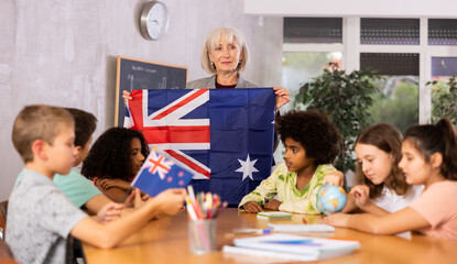 Elderly high teacher, conducting a lesson in the classroom, holds the national flag of Australia...
