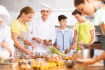 Young guy cook in uniform teaches group of children how to cook dish