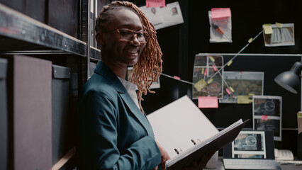 Female police officer reading case information in file, standing in incident room. Private detective using witness statements papers and records to do research, solving criminal activity. - obrazy, fototapety, plakaty