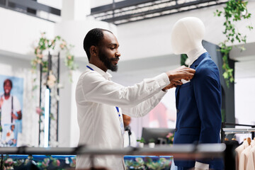 Clothes retail shop african american assistant dressing mannequing in formal suit to showcase apparel in stock. Shopping mall fashion department man employee putting male blue jacket on model