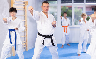 Proficient middle-aged trainer practicing kata standing in row with preteen attendees of karate classes in sports hall