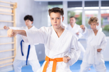 Concentrated teenager in white kimono practicing punches in gym during group martial arts workout....