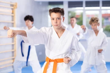 Foto op Aluminium Concentrated teenager in white kimono practicing punches in gym during group martial arts workout. Shadow fight, combat sports training concept .. © JackF