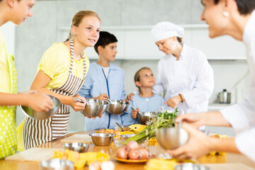 Interested teen girl taking part in cooking classes for children, mixing sauce in bowl with whisk, carefully listening to advices of professional chef instructor