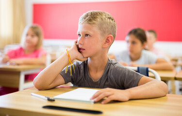 Fototapeta na wymiar Sad bored schoolboy sitting in classroom during lesson in elementary school