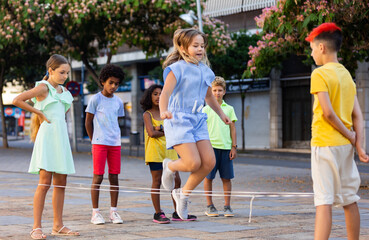 Obraz premium Multiracial group of cheerful preteen children having fun together outdoors on summer day, playing chinese jump rope on city street..