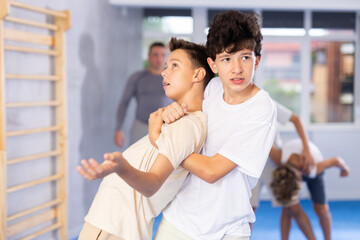 Fototapeta premium Determined teenagers sparring at self-defense training, trying to break free of back grab. Boys practicing basic techniques under guidance of experience instructor....